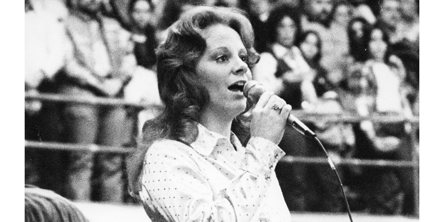 A black and white photo of Reba McEntire singing with the crowd standing up with their hands over their chests behind her.